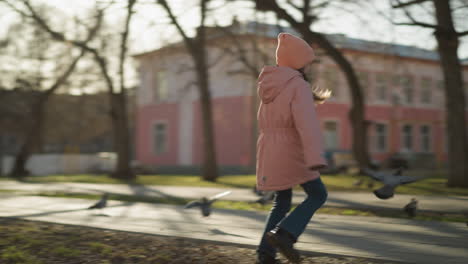 joven alegre en chaqueta rosa y vaqueros azules persiguiendo palomas en un camino de parque soleado, rodeado de árboles y edificios en el fondo