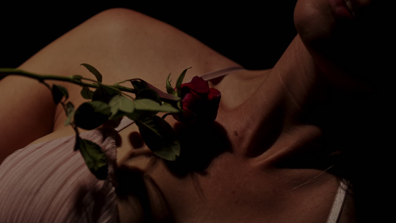 An attractive woman in enticing lingerie sensually strokes a red rose on her beautiful body in slow motion