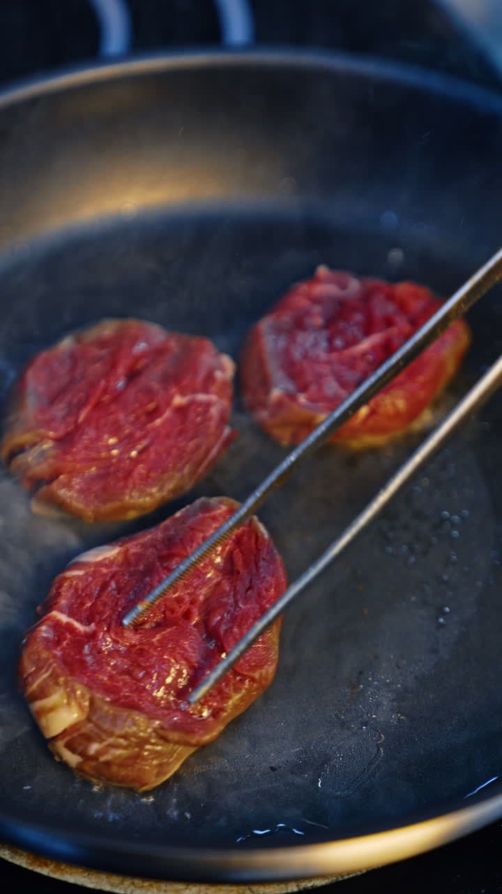 Three pieces of beef are fried in the frying pan. Metal tongs are used to move the meet preventing sticking. Close up. Vertical video.