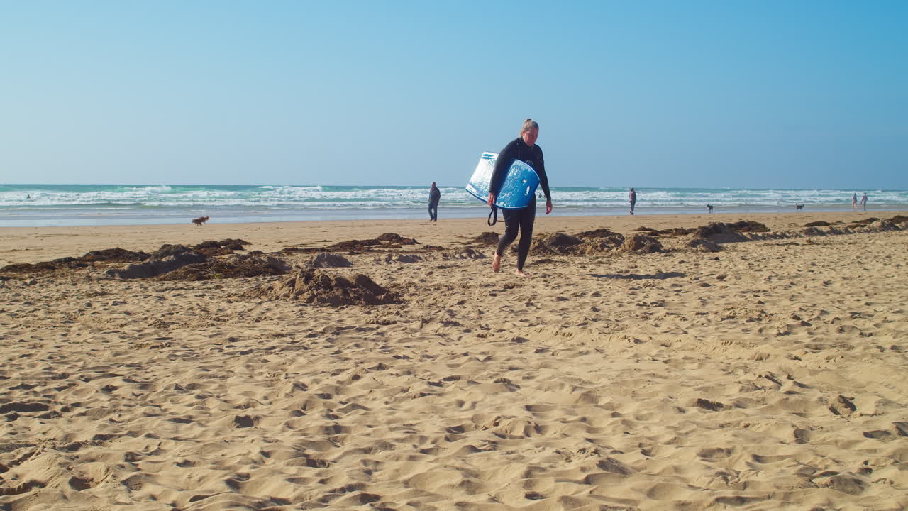 Woman Surfer Walking Back from Surf on Perranporth Beach During Summer In Cornwall, England - Tracking Shot