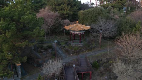 drone aerial view in south korea countryside lowering in front of a asian temple surrounded by green trees and wooden path jeju island