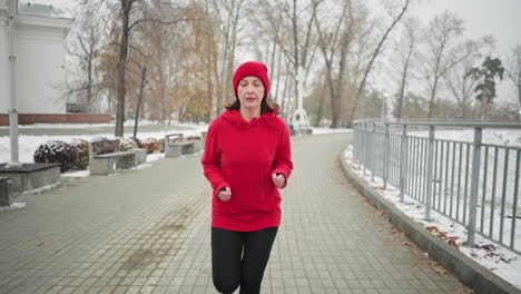 woman jogging looking tired along snowy park pathway in red hoodie and black leggings, surrounded by serene winter scenery with benches, bushes, iron railing, lamp posts, and distant cross monument