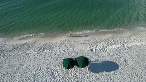 Drohnenansicht-Eines-Mannes,-Der-An-Weißen-Sandstränden-Des-Golfs-Von-Mexiko-Läuft,-Mit-Klarem-Smaragdfresser-Und-Zwei-Grünen-Sonnenschirmen-Am-Strand