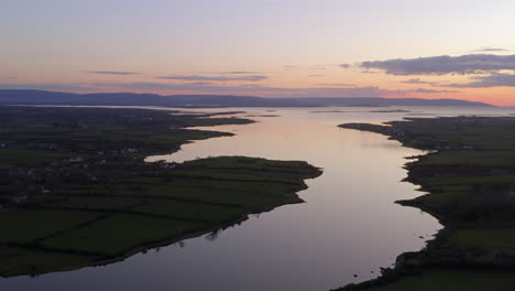aerial pullback orbit of river winding through clarinbridge with sunlit sky and dark landscape contrast. galway, ireland