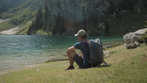 Cámara-Recorriendo-A-Un-Excursionista-Que-Está-Sentado-Junto-A-Un-Lago-De-Montaña-Y-Mirándolo-Con-Admiración