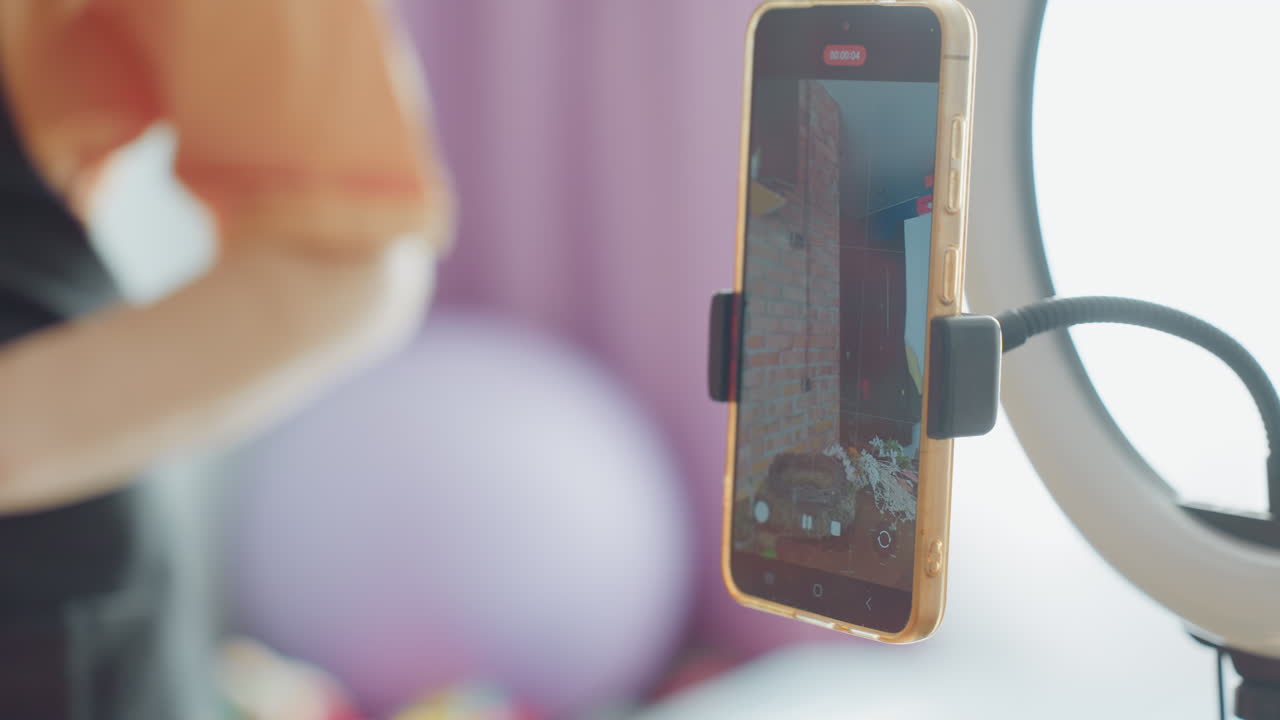 Close up of female hands with red manicure adjusting smartphone on tripod mount in front of ring light, preparing for online recording focusing camera for professional content creation