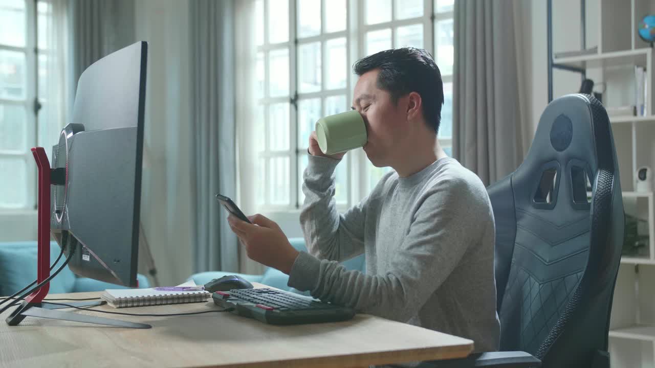 Asian Man In Long Sleeved T-Shirt Using Mobile Phone And Drinking Coffee While Using Desktop Computer For Working At Home.