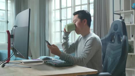 asian man in long sleeved t-shirt using mobile phone and drinking coffee while using desktop computer for working at home.