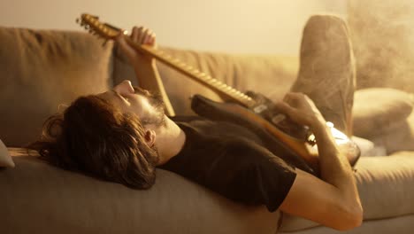 man playing on guitar while lying on couch in smoky room