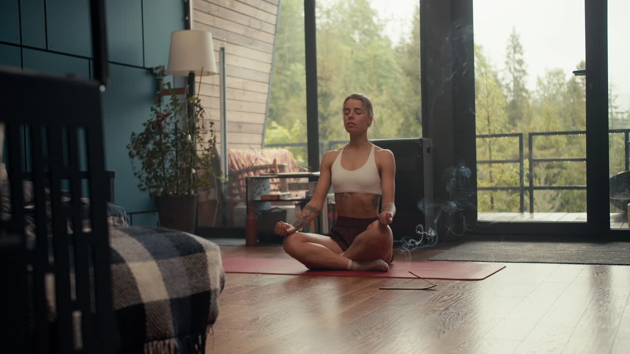 A blonde girl in a white top with tattoos is meditating and using incense that smolders and emits a small amount of smoke. Meditation in a country house overlooking a green coniferous forest