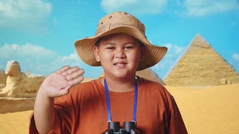 asian boy with hat and binoculars using magnifying glass, smiling and waving hand while traveling in giza pyramid. boy researcher examines something, travel adventure, close up