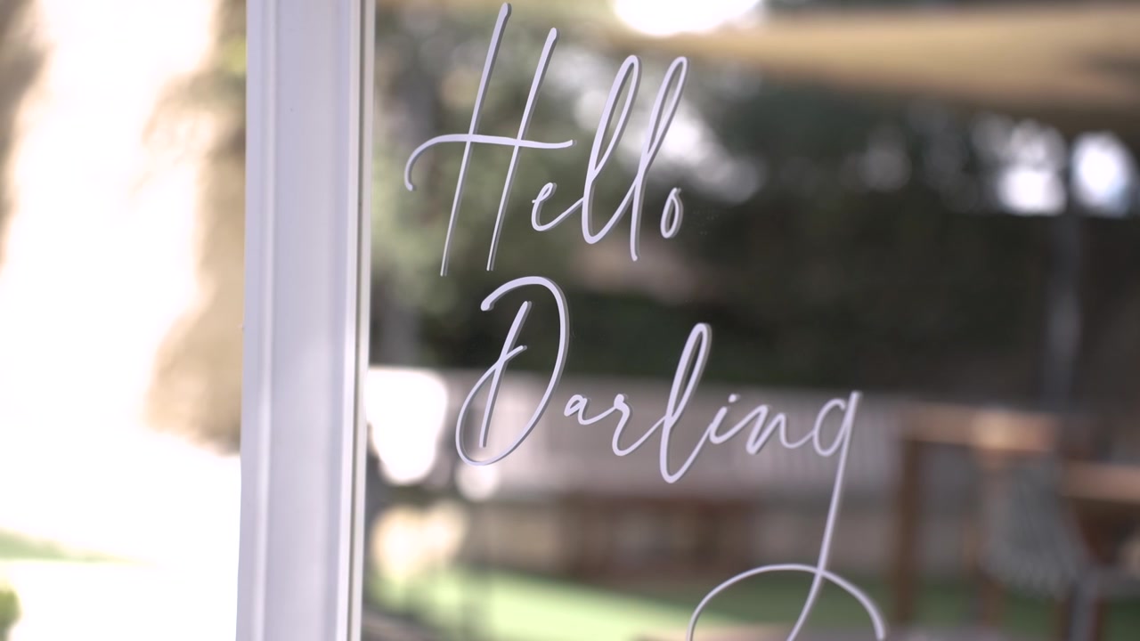 Un'insegna con le parole "Hello Darling" è visibile in un caffè. La location è vicino a un parco. La luce del sole illumina l'area. Si sentono persone parlare in sottofondo.