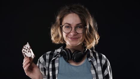 mujer joven morena con gafas y auriculares posando juguetón comer sostener en la mano una barra de chocolate con nueces aislado sobre fondo negro en el estudio. personas emociones sinceras concepto de estilo de vida