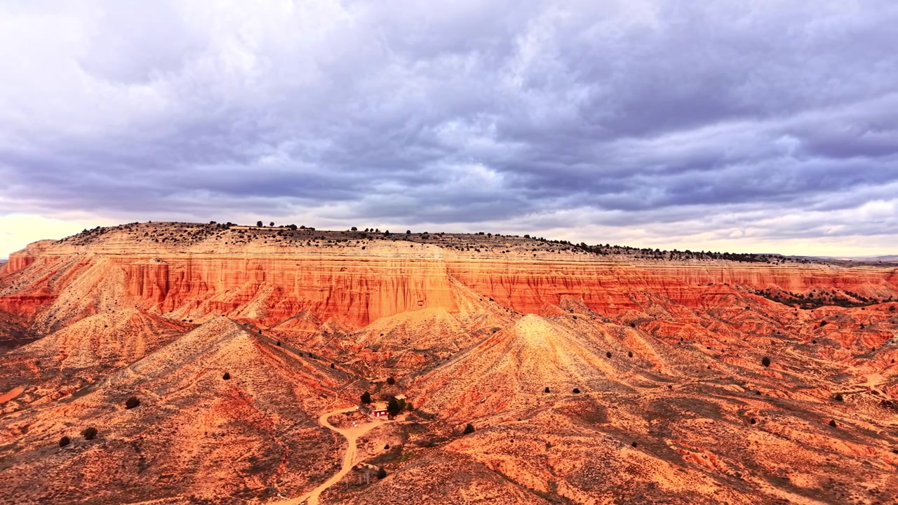 Red sandstone mesa and badland landscape under stormy sky. Wide drone perspective of flat topped cliff face with orange and ochre rock layer, eroded gully, sparse conifer, dirt track.