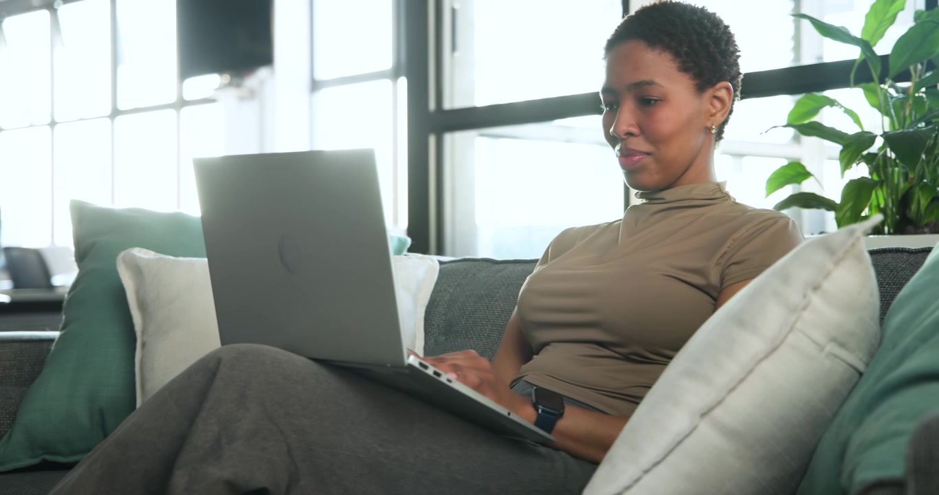 Afroamerikansk kvinde falder til ro i sofaen, skriver på sin bærbare computer, tjekker uret og lukker computeren for at rejse sig.
