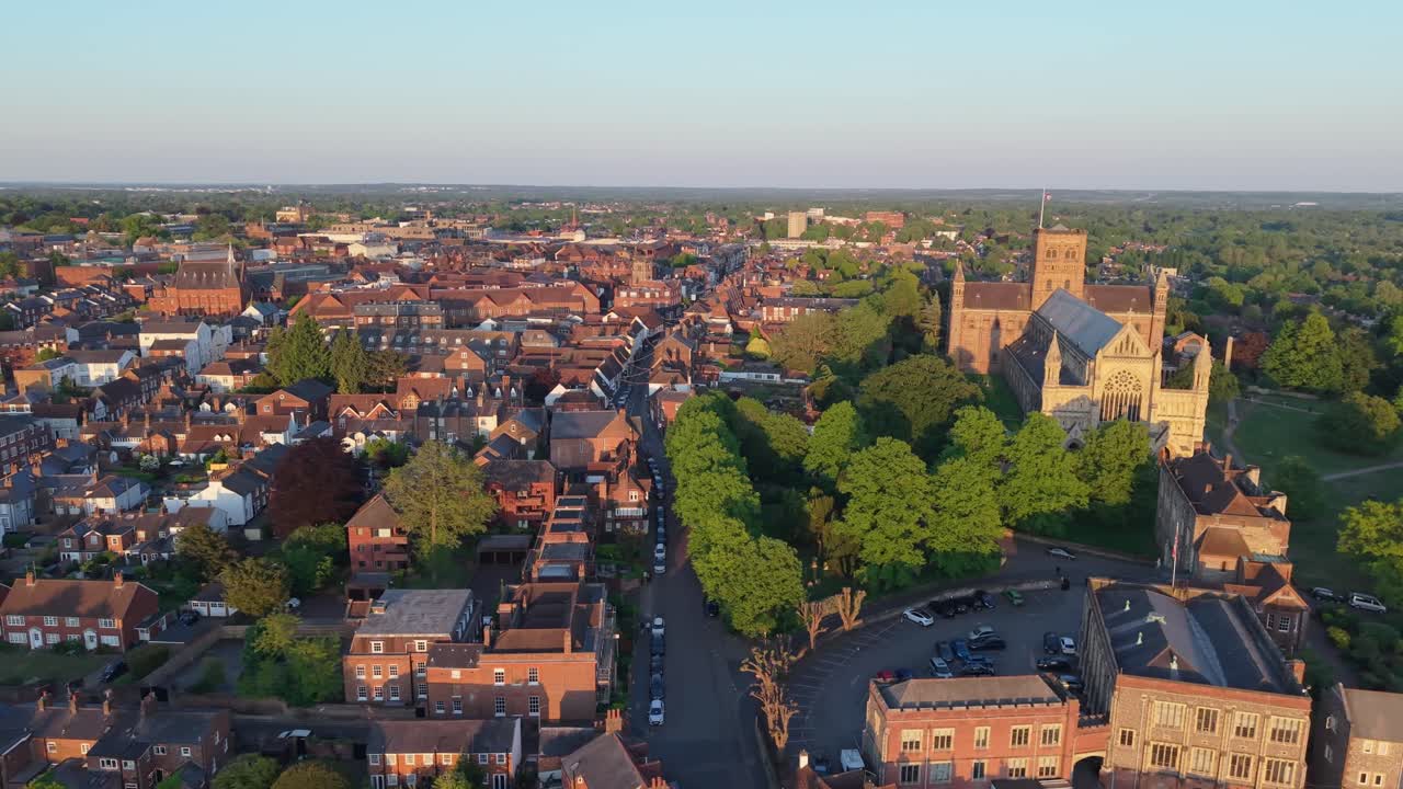 A cinematic parallax drone shot captures the picturesque cityscape of St Albans bathed in warm afternoon sunlight, highlighting historic architecture and charming streets