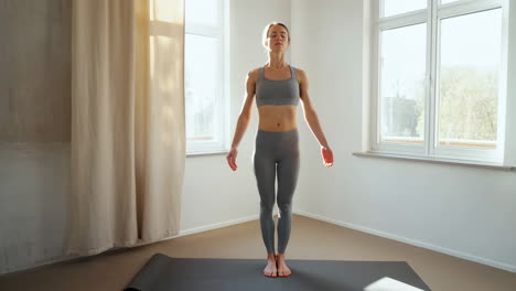 a woman stands in a meditative or preparatory yoga pose on a mat indoors