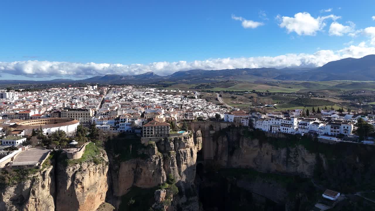 Puente Nuevo bridge gracefully connecting the white houses of Ronda, Spain, spans the deep El Tajo gorge, with majestic mountains rising in the background on a bright, sunny day