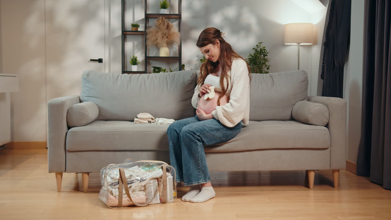 Pregnant woman sitting on sofa preparing baby clothes for hospital bag anticipating childbirth and