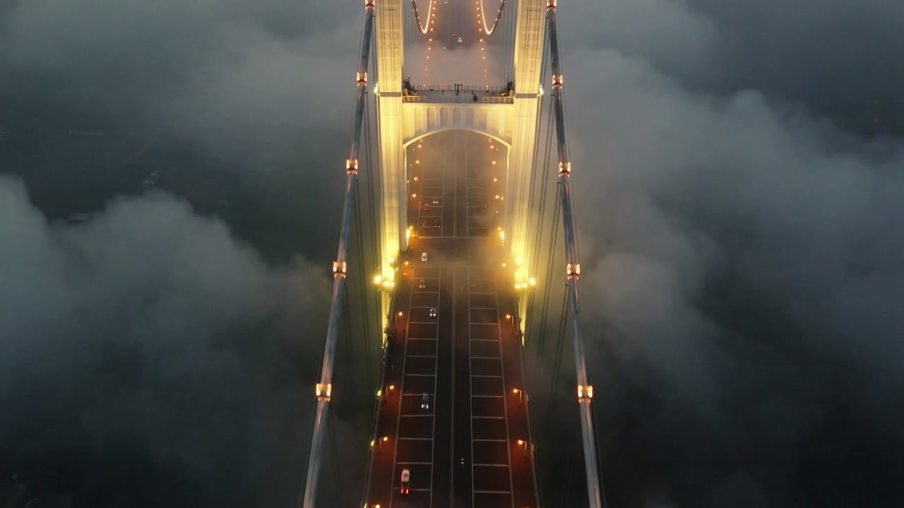 Moving drone revealing central span and lit suspension tower at foggy bridge, headlights appearing