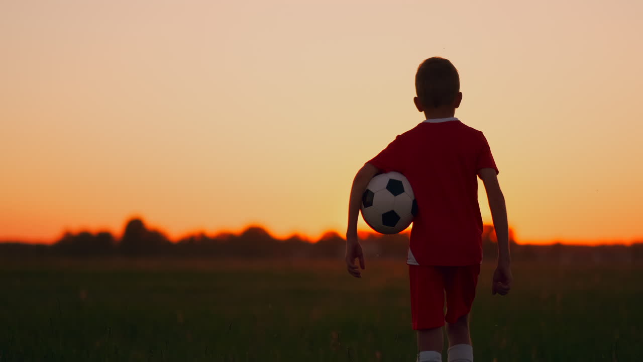 el jugador de fútbol con la pelota atraviesa el campo la cámara de puesta de sol sigue al niño. niño al amanecer viendo el amanecer de pie con una pelota de fútbol.