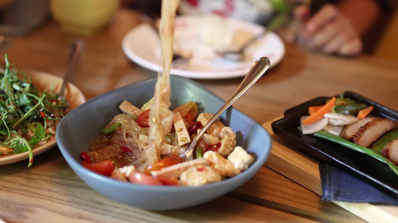 Hand stirs spicy Thai glass noodle salad in bright restaurant, surrounded by fresh side dishes