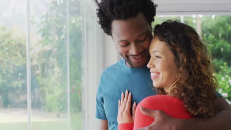 Happy-biracial-couple-embracing,-standing-at-window-and-looking-into-distance