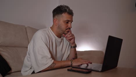 side view of man typing on laptop with serious expression while researching information online in a cozy home setup
