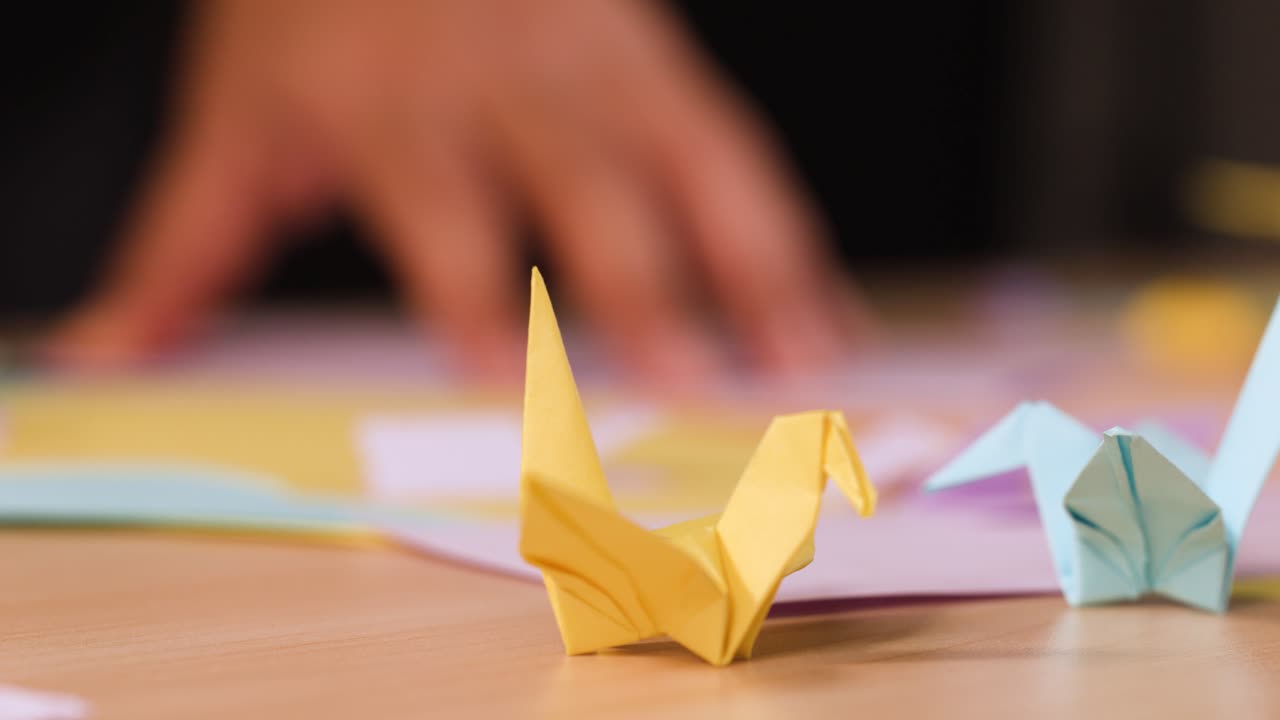 Una gru di carta gialla è appoggiata su un tavolo di legno, mentre delle mani piegano della carta sullo sfondo
