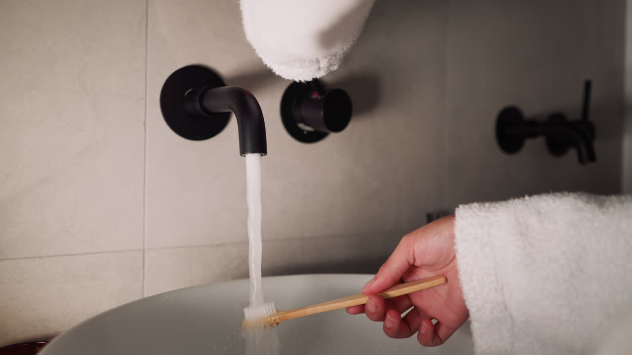 Woman washes toothbrush with running water in bathroom closeup. Lady in soft terry bathrobe cleans brush after teeth washing at home. Personal hygiene