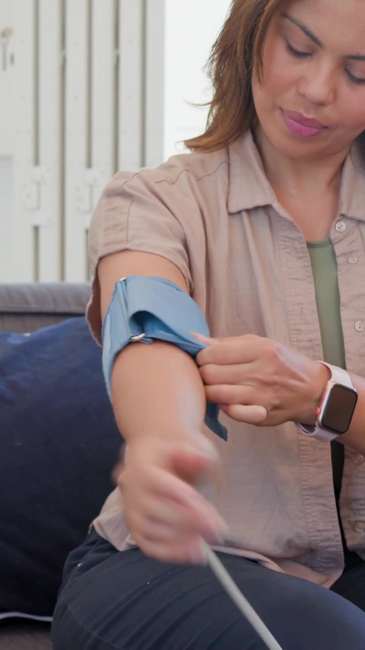 Video verticale: Donna che apre un bracciale blu e se lo infila sul braccio sinistro, sul divano di casa, regolandolo per la misurazione della pressione sanguigna.