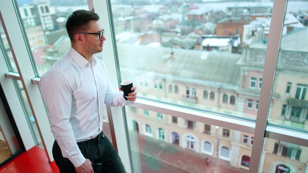 Sportive Caucasian male in white shirt stands at the window looking at the city. Man drinking coffee during work break.