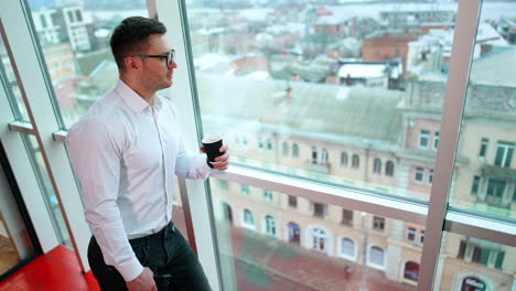 sportive caucasian male in white shirt stands at the window looking at the city. man drinking coffee during work break.