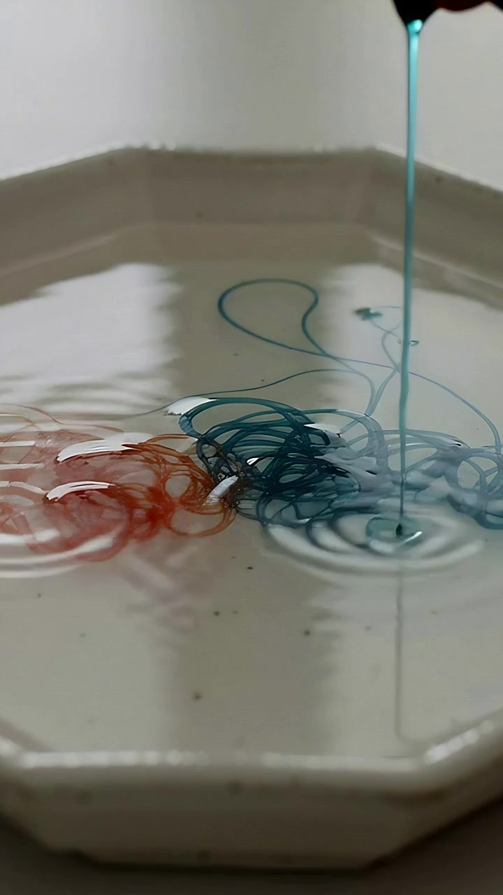 Vertical video: Descending orange and teal streams forming coils on off-white dish, creating knots