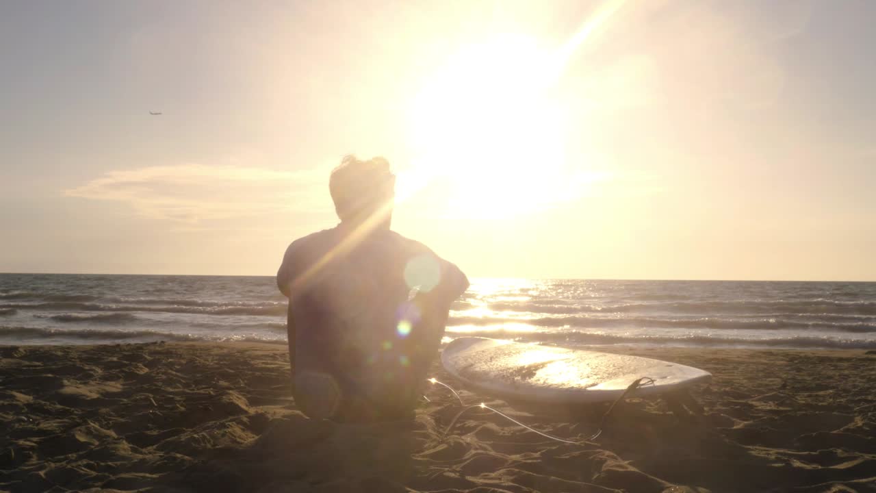 해변에서 모래에 누워있는 서핑 보드와 함께 앉아있는 젊은 남성 서퍼가 해가 지는 바다를 바라보며 음악을 듣고 춤을 추고 있습니다.