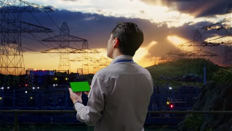 back view of an asian male professional worker standing near high voltage tower, industrial facility, checking on smartphone with green screen and look at the factory once