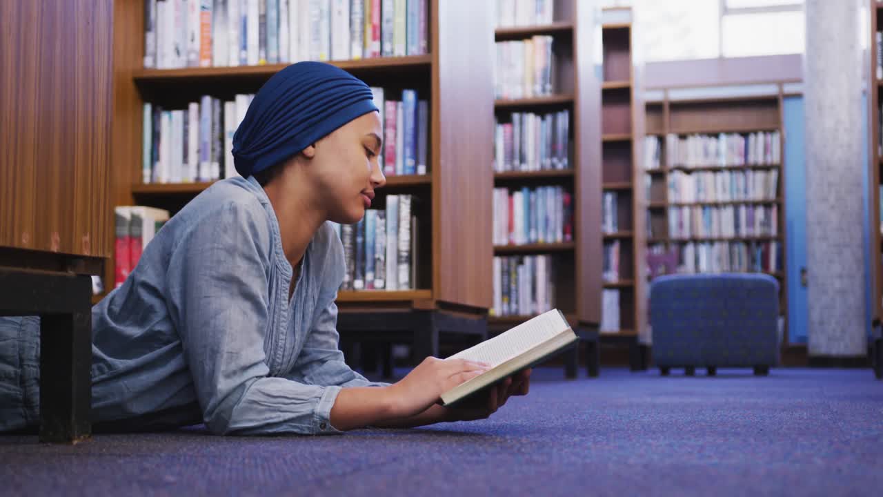 Studentessa asiatica con un hijab blu sdraiata per terra che legge un libro.