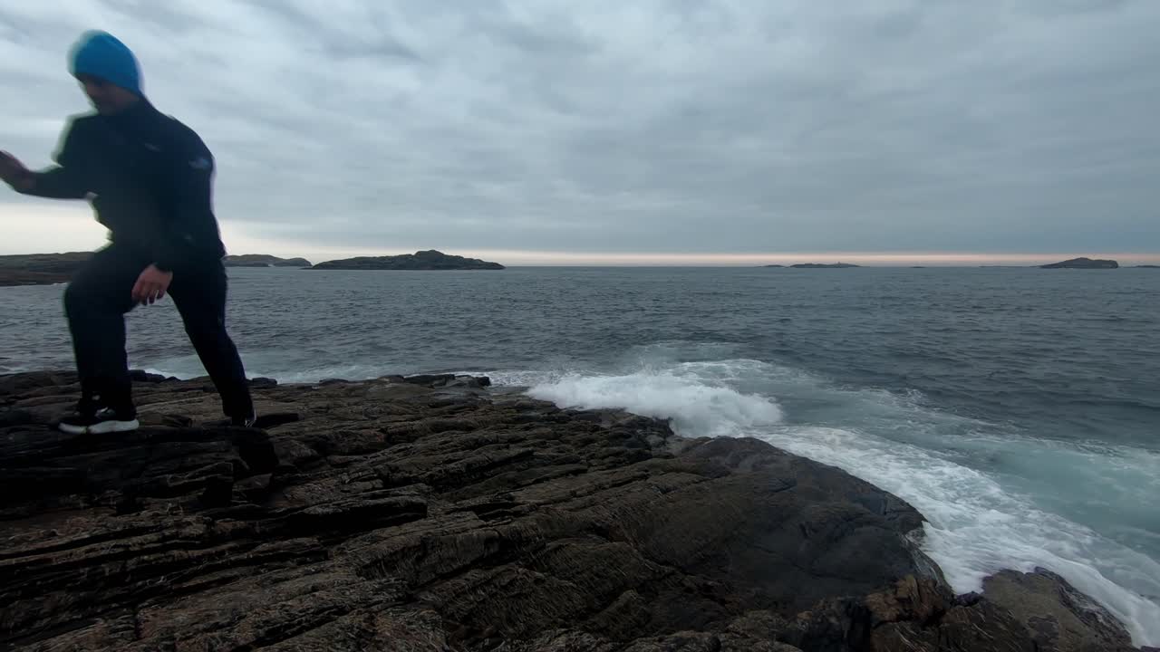adulto masculino sentado en una roca cerca del mar y usando su teléfono celular antes de levantarse y alejarse - clip estático con olas lavándose en la orilla y nubes dramáticas después del atardecer