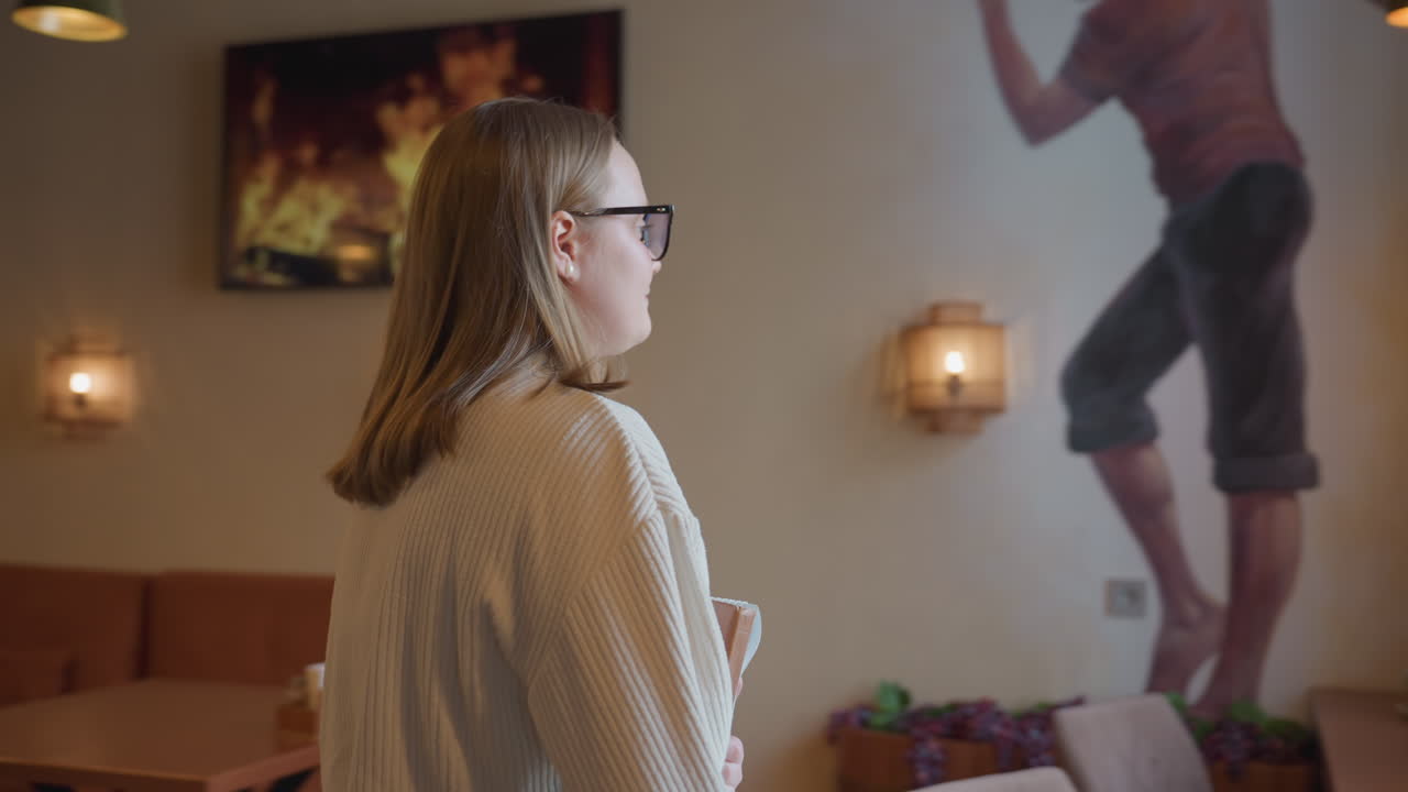 student in white sweater walks into restaurant interior with cozy lighting and plants, holding notebook as she heads toward table where another person is seated, ready to sit and place her book down