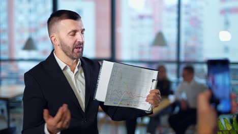 businessman holding a flipchart with graphs and describing them. bearded man speaking emotionally in front of camera. close up portrait.