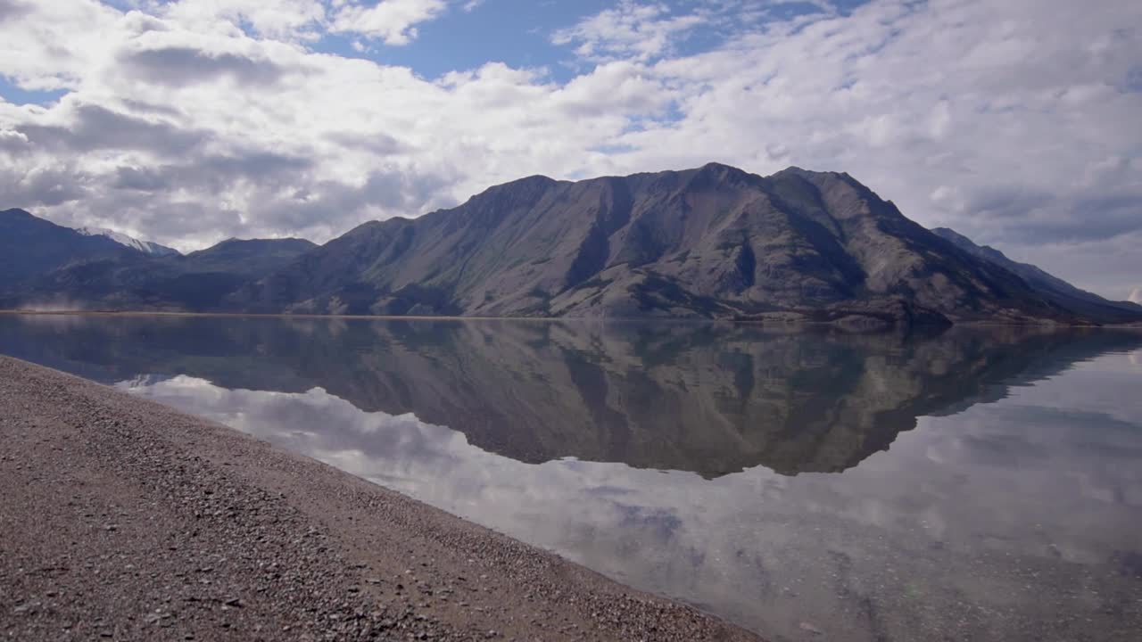 paisaje salvaje escena de verano del lago kluane espejo brillante de yukón por limo marrón, suciedad y piedras por el borde del lago y la impresionante montaña de ovejas en el fondo en el día soleado del cielo azul, antena ascendente