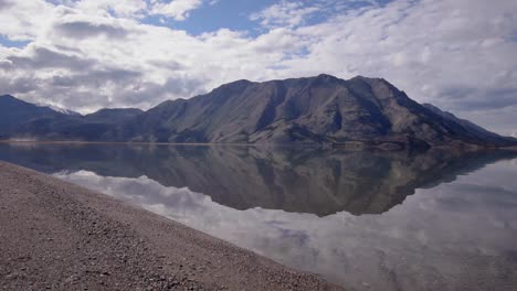 Wilderness-landscape-summertime-scene-of-Yukon-shiny-mirror-Kluane-lake-by-brown-silt,-dirt-and-stones-by-lakeside-edge-and-stunning-Sheep-mountain-in-background-on-sunny-blue-sky-day,-rising-aerial