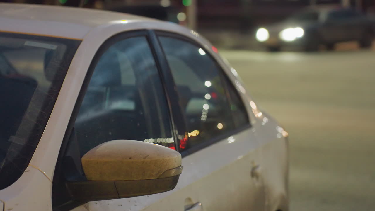 Parked white car on roadside during night with blurred reflections of moving cars and pedestrians, glowing street lights and headlights creating soft atmospheric urban scene with motion and contrast