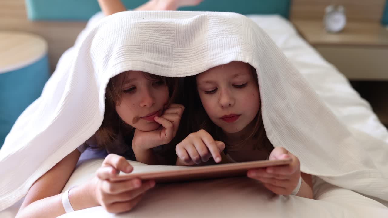 Two children using a tablet under a blanket