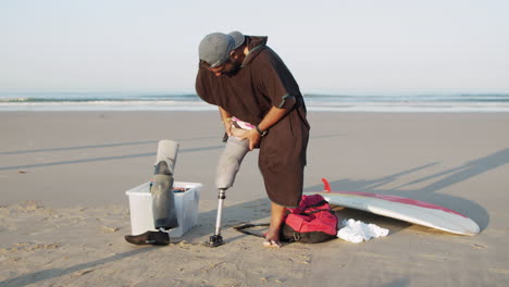 long shot of a male surfer on the beach putting prosthetic leg on