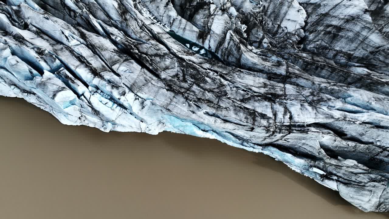 Vista cenital con dron del borde de un glaciar blanco azulado con vetas de morrena oscura que se une a un lago de agua de deshielo color canela, creando una llamativa frontera natural