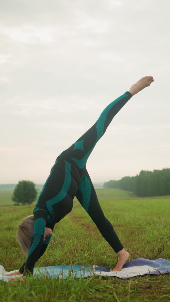 mujer en traje verde y negro practicando postura de yoga dividida de pie al aire libre en una alfombra de yoga en un campo cubierto de hierba bajo un cielo nublado, estirando su cuerpo hacia abajo como parte de una rutina de bienestar