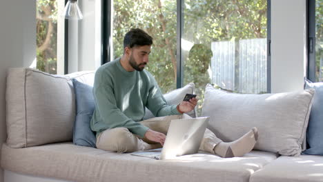 biracial man sitting on sofa using laptop for online shopping at home, slow motion