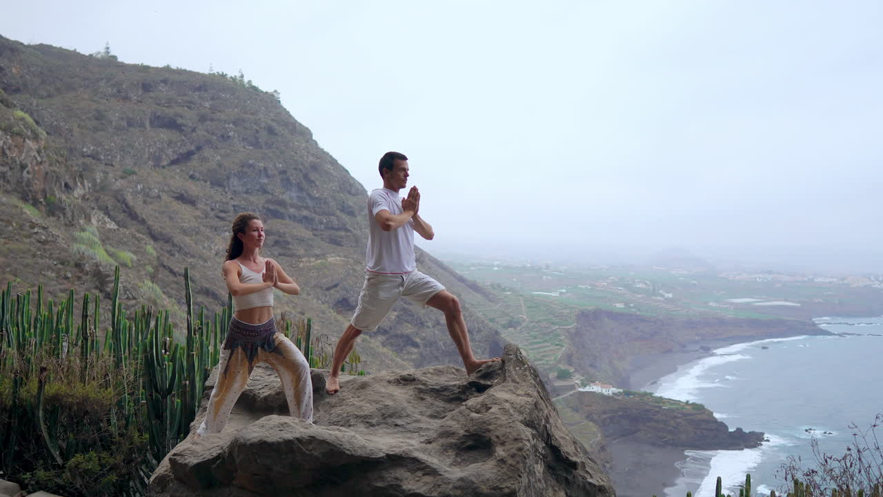 On a cliff, a man and woman lift their hands, breathing in the sea breeze during yoga while overlooking the ocean's expanse