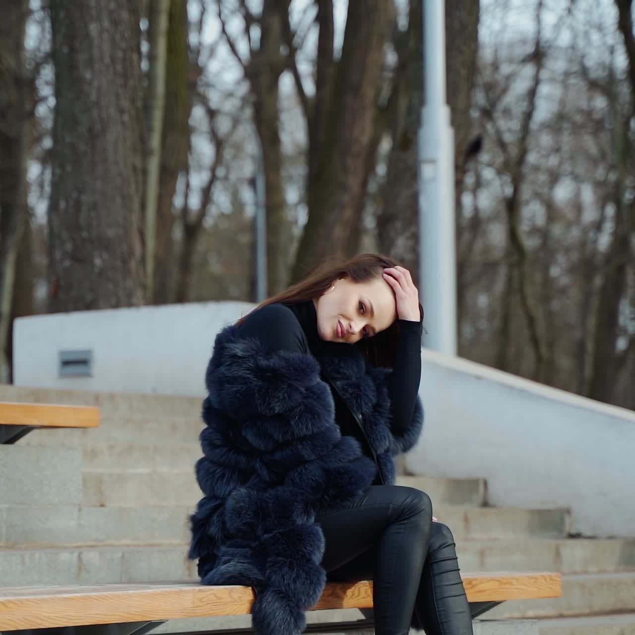 Luxury girl sitting on bench. Attractive young woman in fur coat enjoys fresh air in the park and smiles. Positive brunette outdoors.